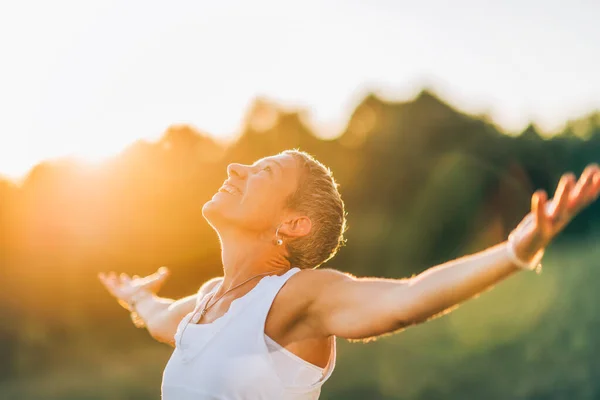Energized and happy female with the sun shining behind her outside in a field with outstreched arms.